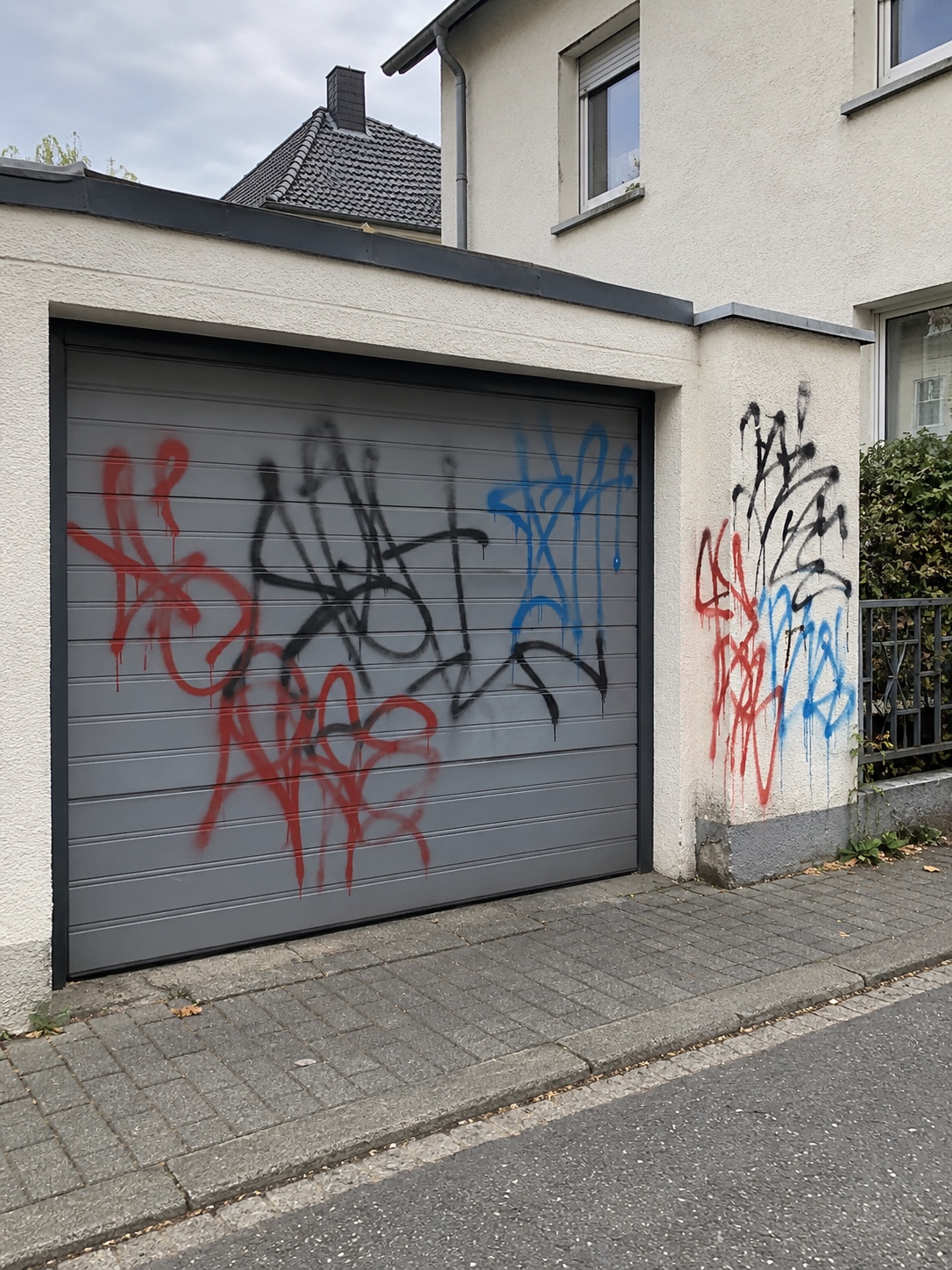 Rote, schwarze und blaue Graffiti bedecken das graue Garagentor und die angrenzende helle Putzwand neben dem Wohnhaus